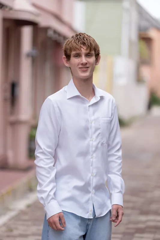 Spencer wears a white button-up shirt and light jeans while standing alone on a brick street lined with pastel-colored