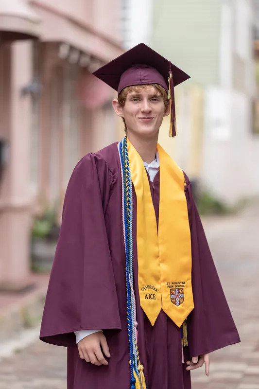 Spencer wears his purple graduation gown and golden honors cords on a brick-lined street in Downtown St Augustine.