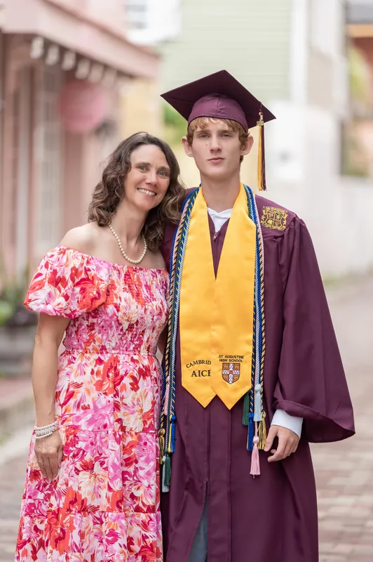 A woman in a pink floral dress smiles beside Spencer in his cap, gown, and honors cords on a tree-lined street.