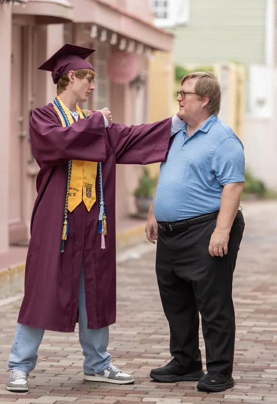 Spencer in maroon graduation robes stands on a brick street in Downtown St Augustine, chatting with a man in a blue shirt.