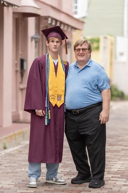 Spencer, wearing his maroon cap and gown with gold honors cords, stands beside the same man on the same brick street.