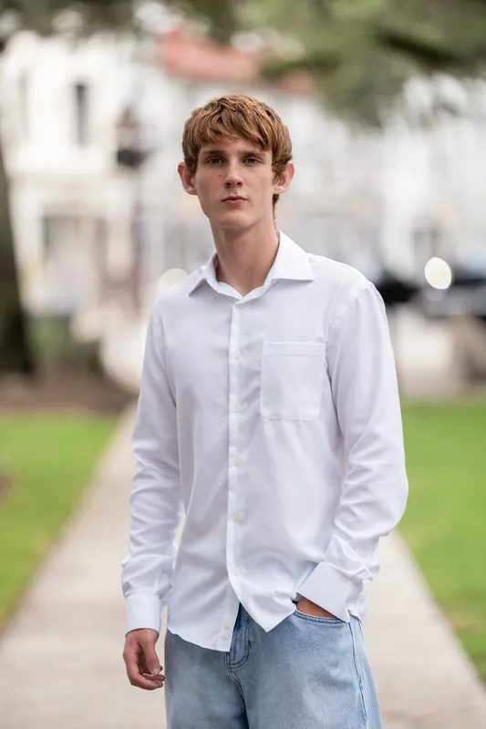 Spencer walks down a tree-lined sidewalk in Downtown St Augustine wearing a white button-up shirt and jeans.