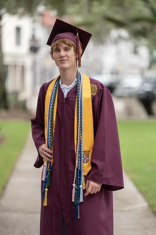 Spencer faces the camera on a tree-shaded street, wearing his maroon cap and gown with gold and teal honors cords draped