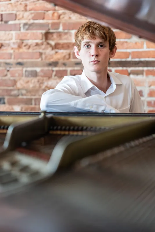Spencer in a white shirt sits at a piano against exposed red brick.