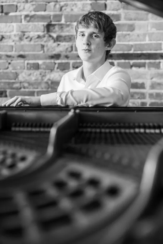 Spencer leans against a piano in profile, rendered in moody black and white.