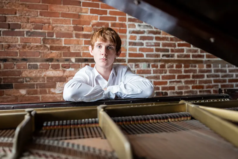 Spencer sits at an open piano with exposed strings, positioned against exposed brick.