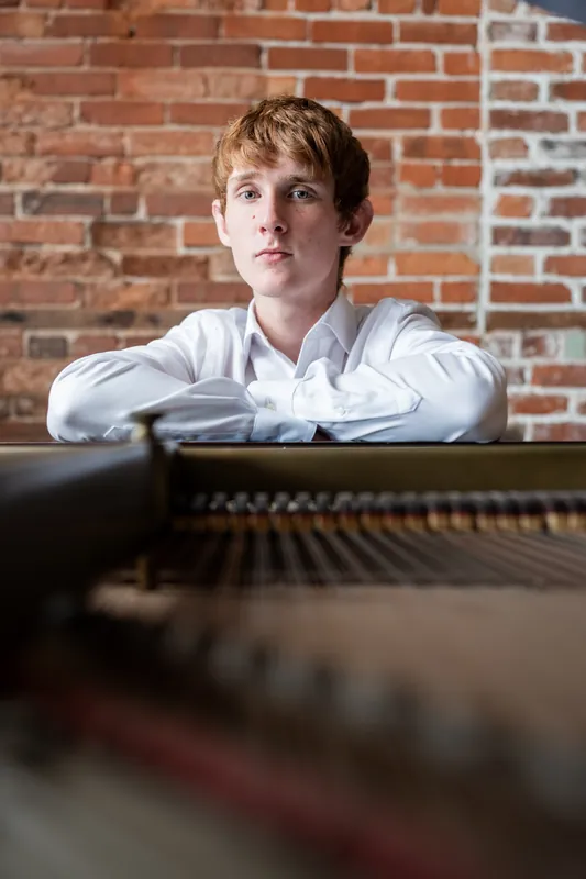 Spencer rests his arms on the piano while gazing directly at the camera.