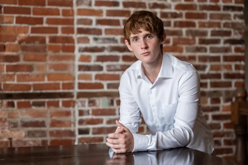 Spencer in a white long-sleeve shirt gazes at the camera with one hand clasped in front of him against brick in Downtown St