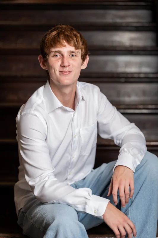Spencer sits on dark steps in a white shirt and jeans, smiling warmly toward the camera in Downtown St Augustine.
