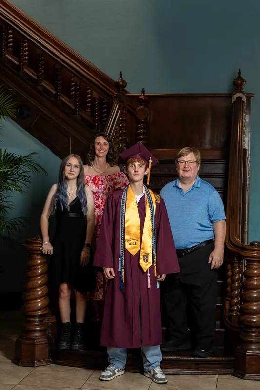 Spencer's family of four stands together on ornate wooden stairs: Spencer in maroon, his mother in black, his father in blue