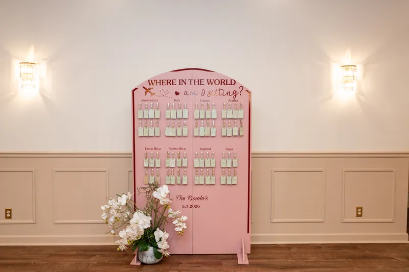Pink seating chart with "Where in the World" theme and white floral arrangement below, flanked by wall sconces