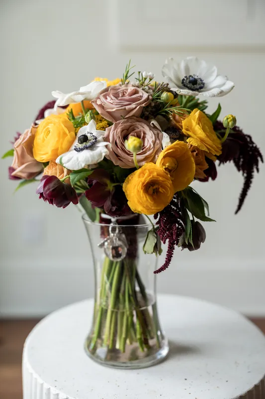 Bridal bouquet with yellow ranunculus, mauve roses, white flowers, and dark burgundy blooms in a clear glass vase
