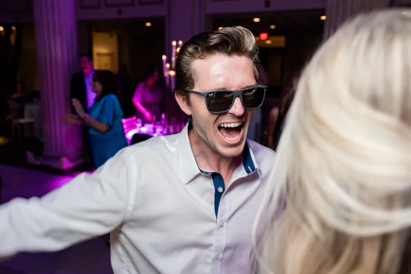Dean laughs while dancing, wearing sunglasses and a light blue shirt as purple lighting fills the reception hall.