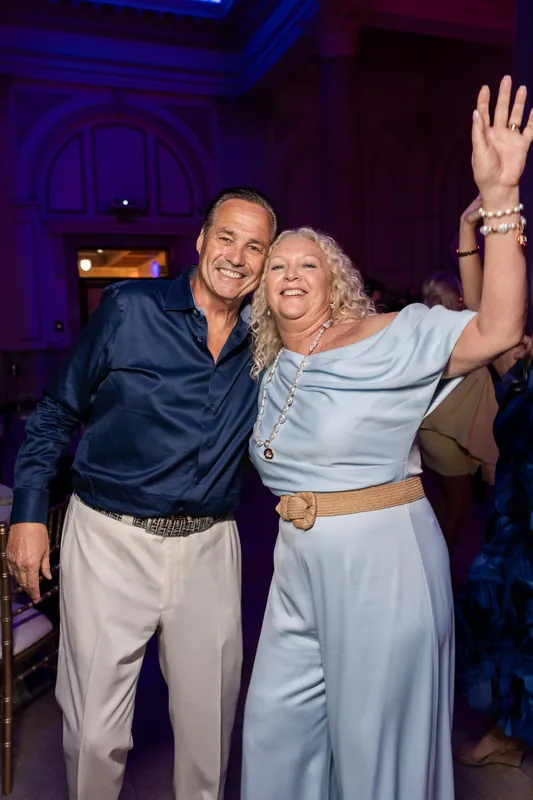 A man in navy and a woman in light blue raise their arms joyfully while dancing under moody ambient lighting.