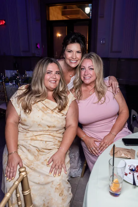 Three women smile together at a table during the reception, dressed in cream, black, and pink.