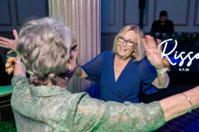 An older woman in glasses reaches toward a woman in a blue blazer as they dance together.