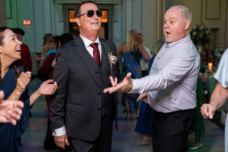 Three men including one in sunglasses and a burgundy tie dance together on the reception floor.