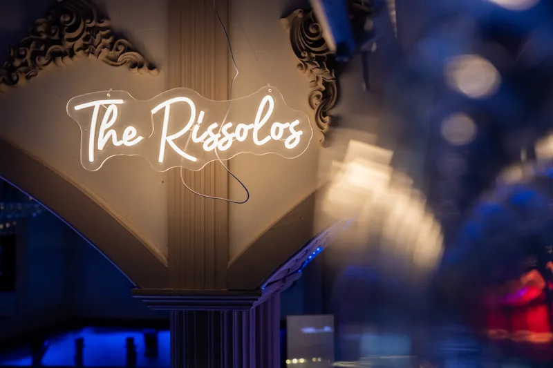 A neon sign reading "The Rissolos" glows above the dance floor with blue lighting in the background.
