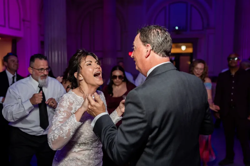 Stacy laughs while dancing with Dean under purple lighting surrounded by guests.