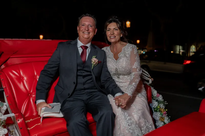 Dean and Stacy sit on a bright red vintage car at The Treasury on the Plaza, dressed in formal wedding attire