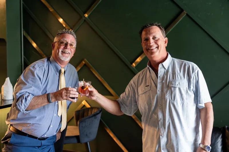 Two men clink beer bottles in a friendly gesture against a geometric green wall.