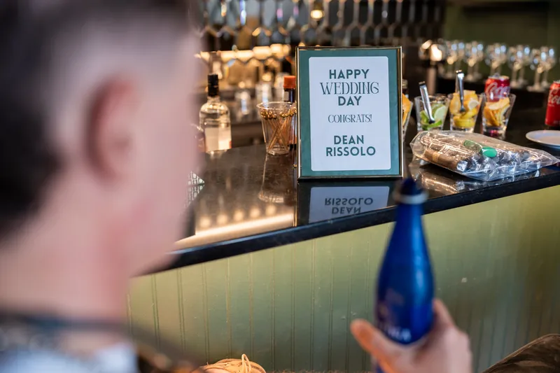 A sign reading "Happy Weddings Day Stacy + Dean Biscolo" sits on a bar counter with glowing bottles behind it.