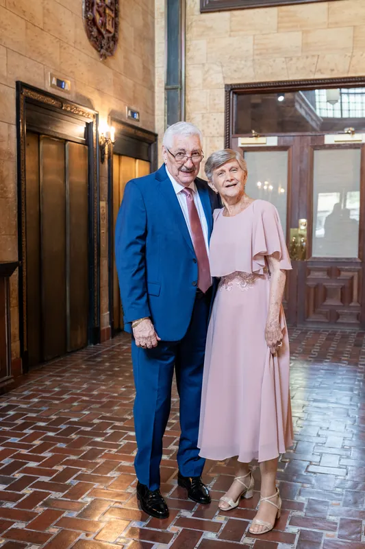 Two guests in formal attire—one wearing a blue suit and red tie, the other in a pale pink dress—stand together in an ornate