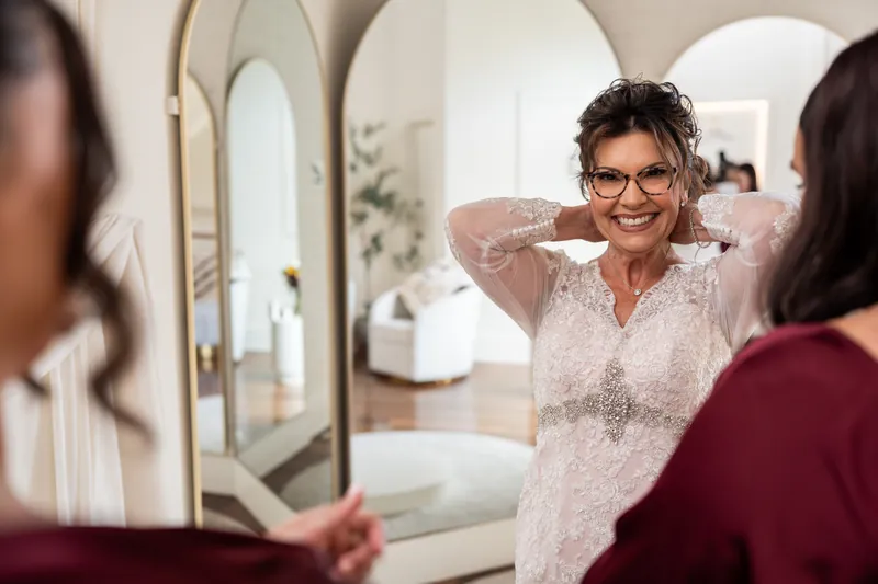 Woman in a beaded ivory lace dress adjusts her hair while looking in a mirror, with another guest visible beside her.