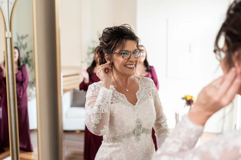 Stacy smiles in a beaded white lace gown with a jeweled sash, adjusting her hair as attendants move around her getting ready.
