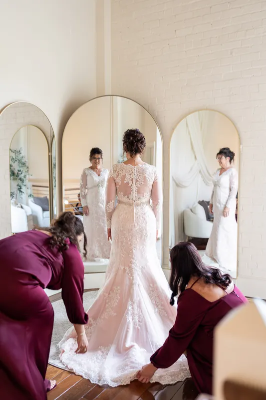 Stacy stands in her beaded white lace wedding dress with a long train as attendants in burgundy adjust the gown's details