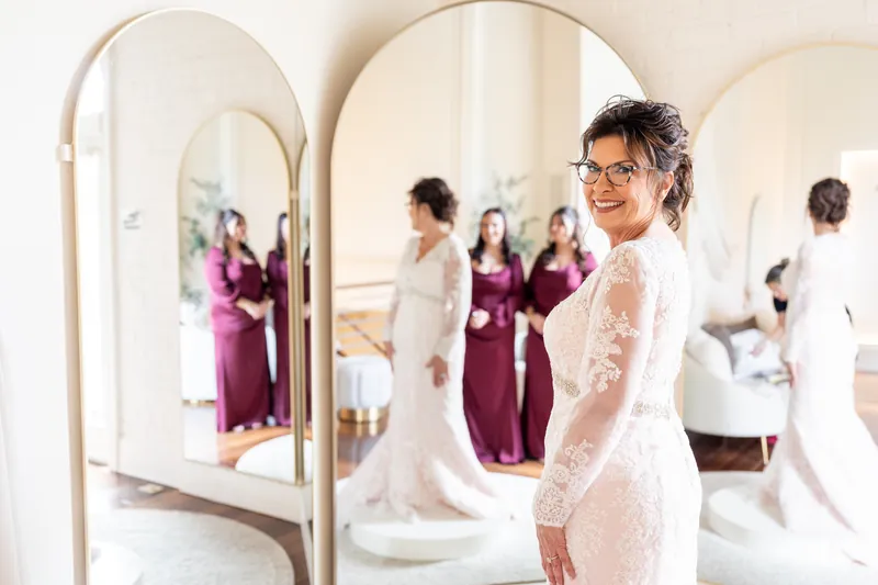 Stacy admires her lace wedding dress in a trio of arched mirrors at The Treasury on the Plaza, surrounded by attendants