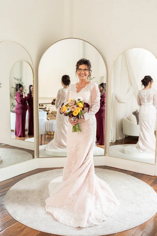 Stacy holds a vibrant bouquet featuring yellow roses, white anemones, and burgundy dahlias while standing before mirrored