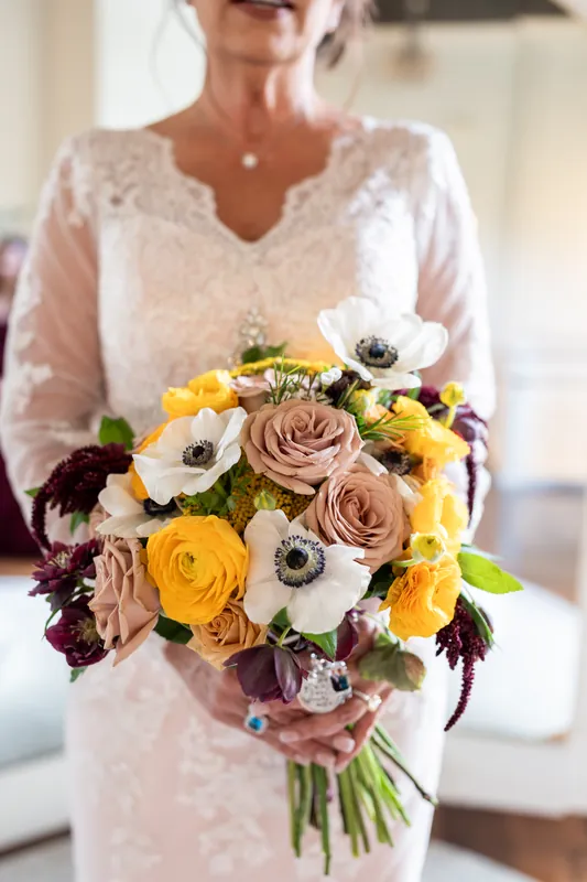 Stacy displays her bridal bouquet of yellow roses, white anemones, taupe roses, and dark burgundy flowers against her lace