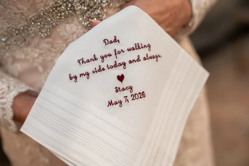 A white handkerchief embroidered with "Thank you for walking by my side and always. Stacy May 11 2026" rests in someone's
