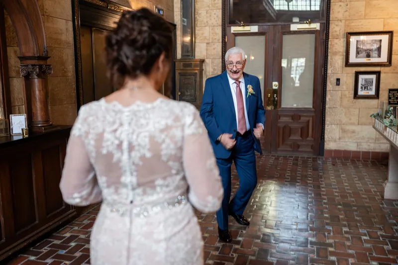 Dean walks toward Stacy down the hallway of The Treasury on the Plaza in a navy suit as she faces away in her lace dress.