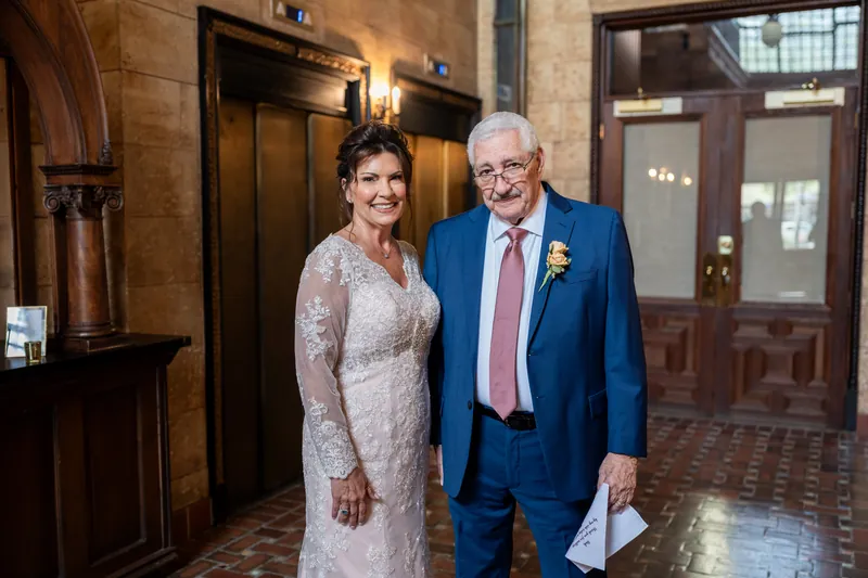 Stacy and Dean stand together in an elegant historic interior, she in a lace gown and he in a blue suit with boutonniere.