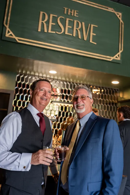 Dean and a guest chat by The Reserve sign, both holding drinks in the venue's contemporary space.