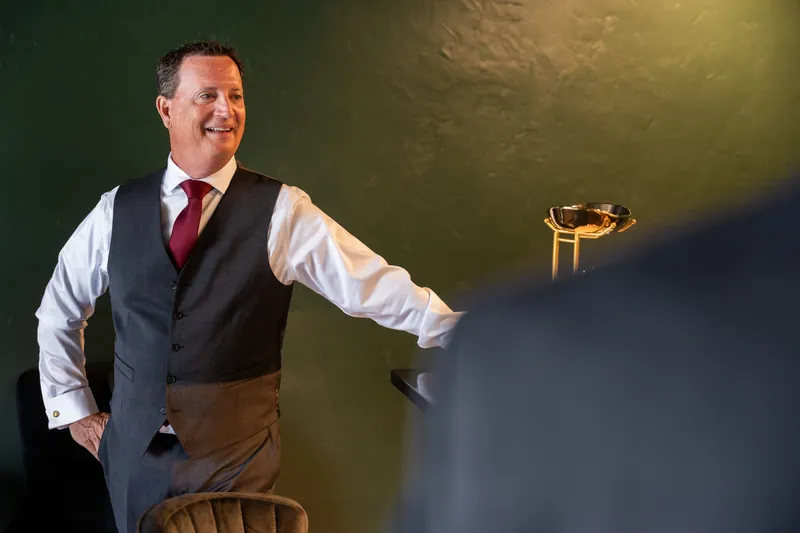 Dean gestures expressively while speaking, wearing a burgundy tie and vest in the softly lit bar.