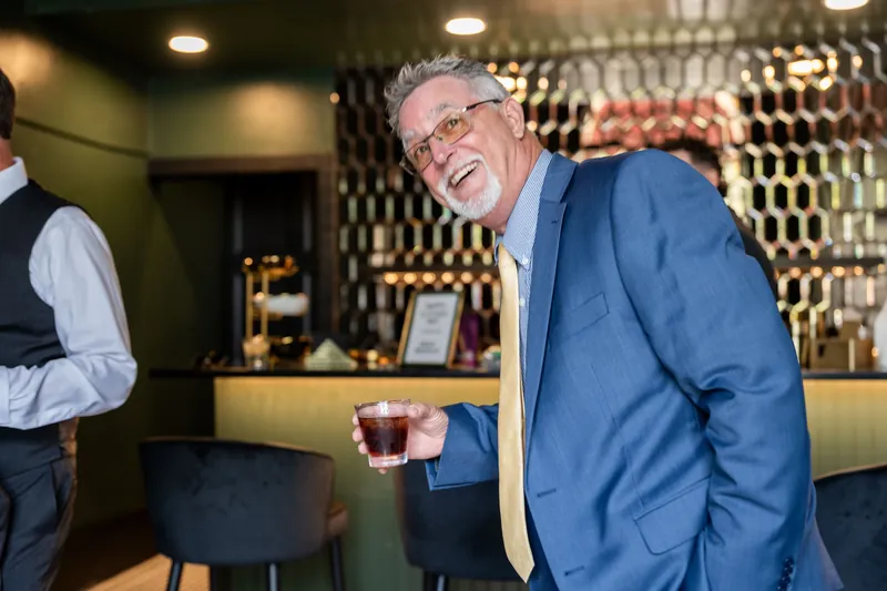 An older man in a blue jacket smiles while holding a drink near the bar's geometric wall pattern.