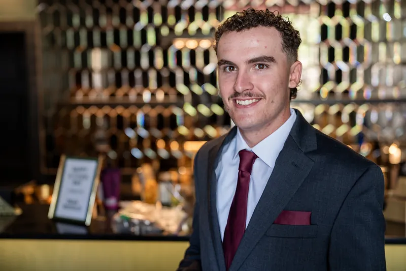 A younger man in a dark suit and burgundy tie smiles at the camera in The Reserve's lounge area.