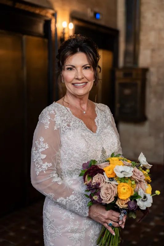 Stacy stands indoors in a beaded ivory gown, holding a yellow and mauve bouquet under warm sconces at The Treasury