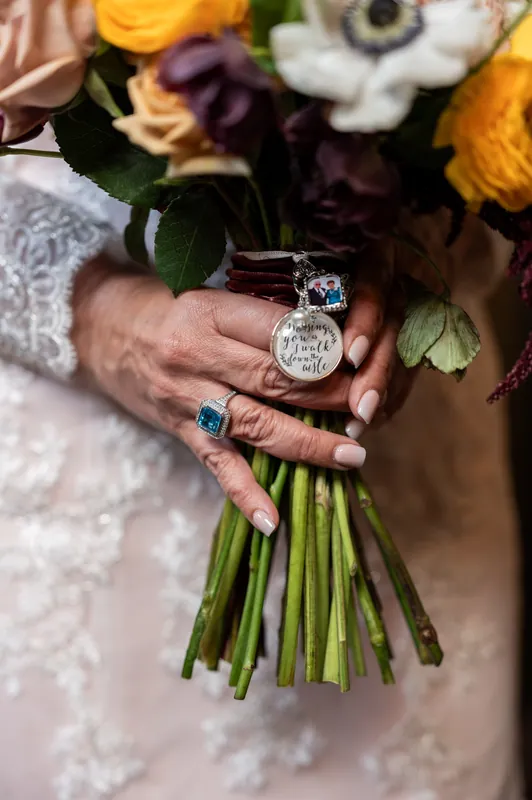 Hands holding a yellow and white bouquet display a silver locket and emerald ring against lace fabric.