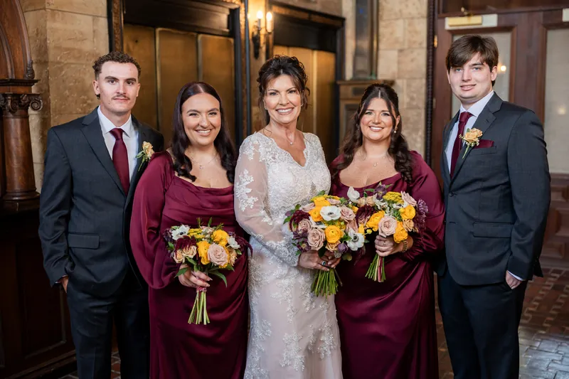 Stacy stands center in lace with Dean and four attendants holding yellow bouquets in burgundy at The Treasury on the Plaza.