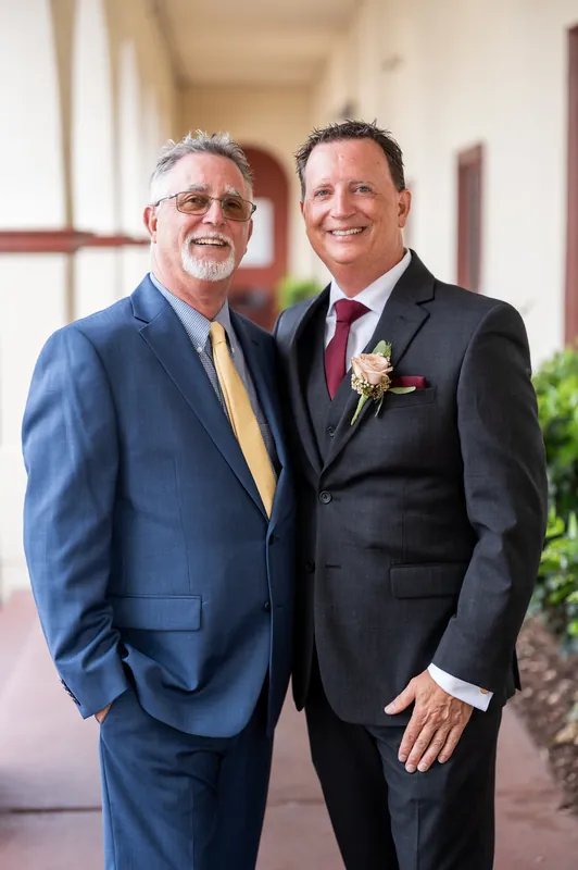 Two men smile together in suits with burgundy ties and floral pins in the venue's elegant hallway.