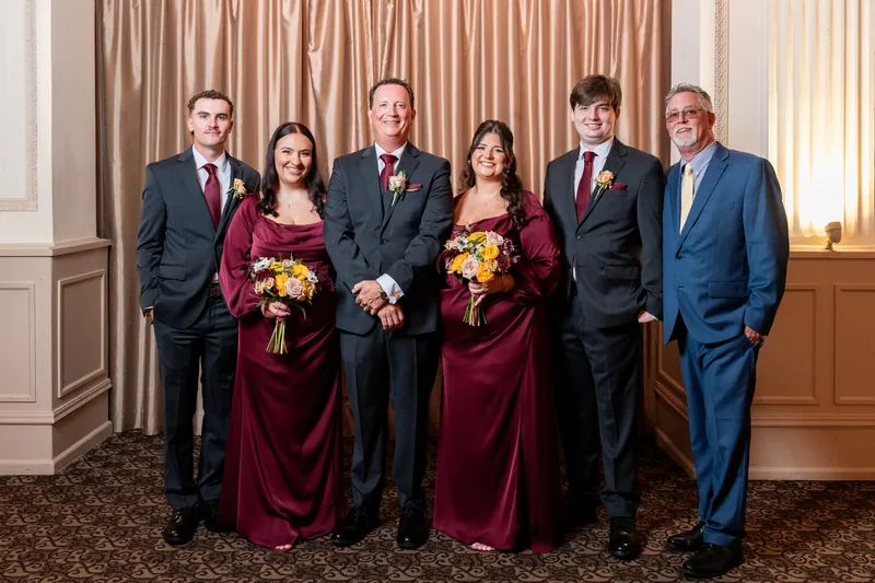 Six people in formal attire stand posed indoors with two holding yellow and burgundy bouquets between draped curtains.