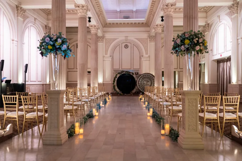 A grand ceremony space features tall columns, floral arrangements, candlelit aisle, and wooden chairs leading to the band