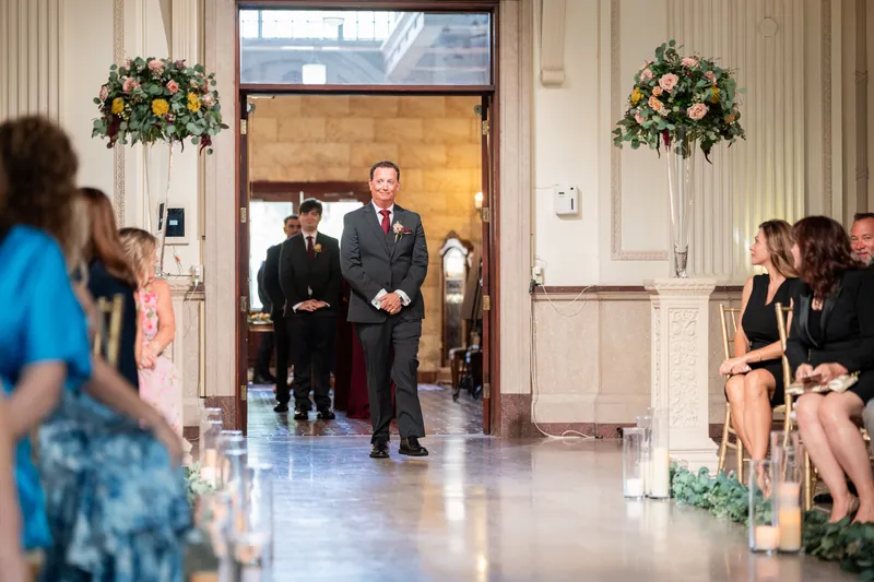 Dean stands at the altar in a charcoal suit as guests watch from the aisles during the processional at The Treasury
