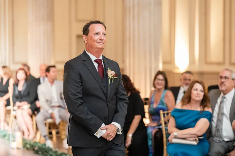Dean in a dark suit and burgundy tie stands with hands clasped, watching as guests settle into their seats.