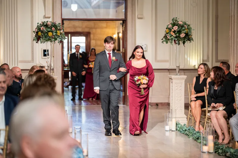 Stacy walks down the aisle in a white gown on the arm of a family member, past seated guests lining both sides.