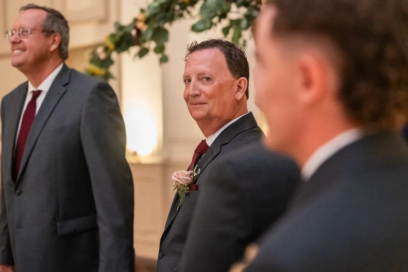 Three men in dark suits and boutonnières stand together in conversation during the ceremony.